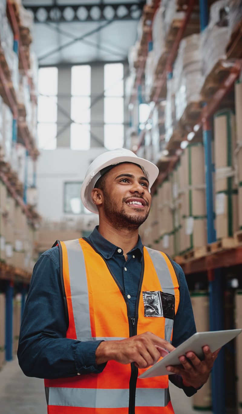 Portrait of young man wearing safety jacket holding digital tablet standing in factory warehouse
