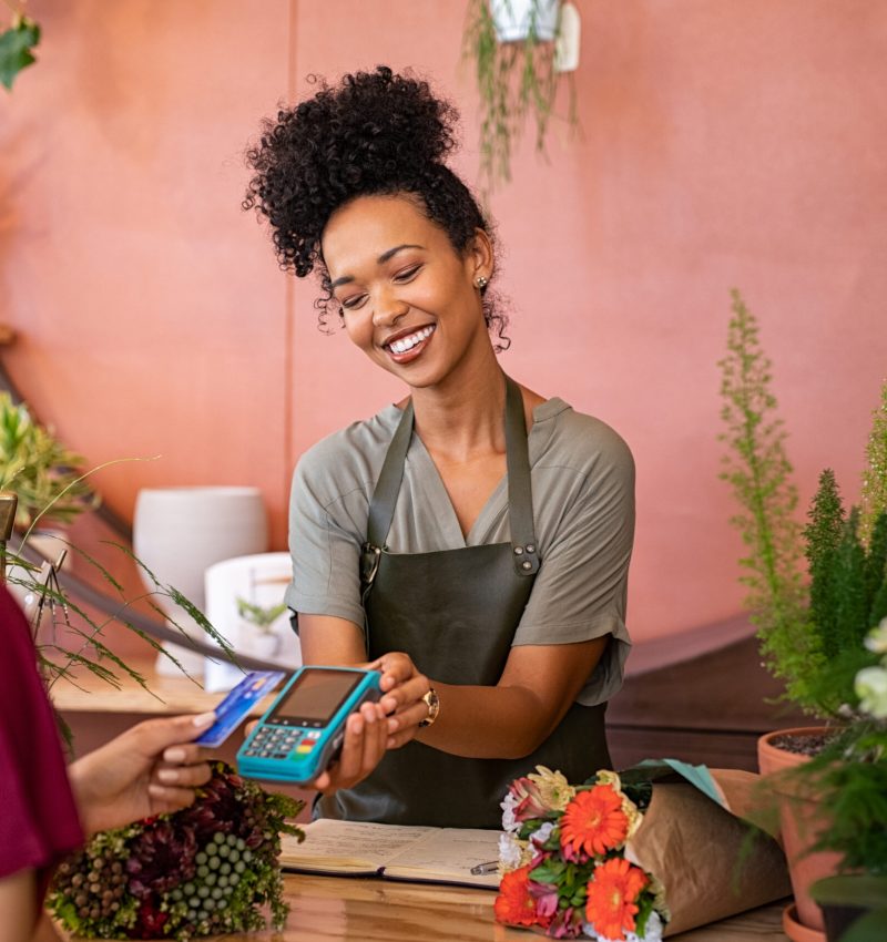 Smiling and friendly florist holding card reader machine at counter with customer paying with credit card. Young african american florist shop assistant holding payment machine while buyer purchase a bunch flower. Woman using bank credit card to make payment by NFC machine.