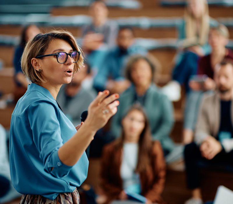 Mid adult businesswoman talking to group of seminar attendees in conference hall.