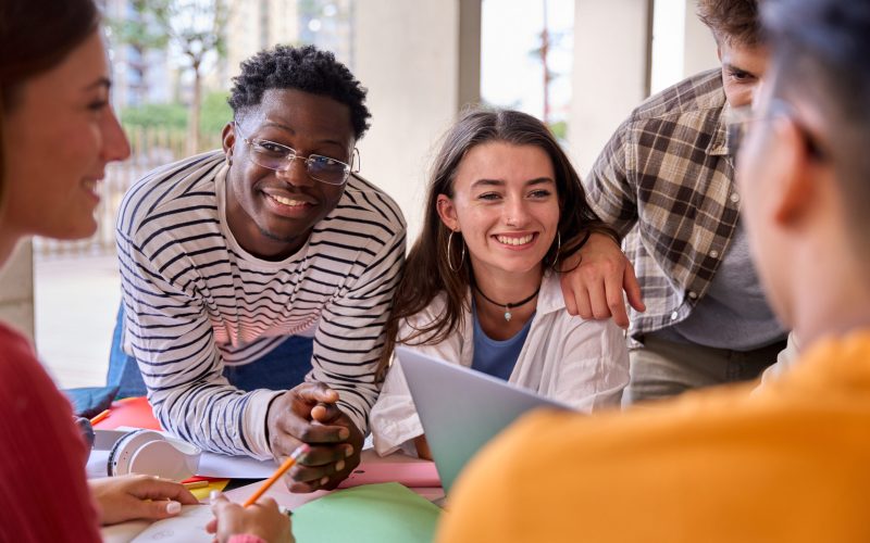Cheerful multiracial couple of young students working together with their classmates on a project sitting at table in faculty. Group of smiling university diverse generation z gathered for class work